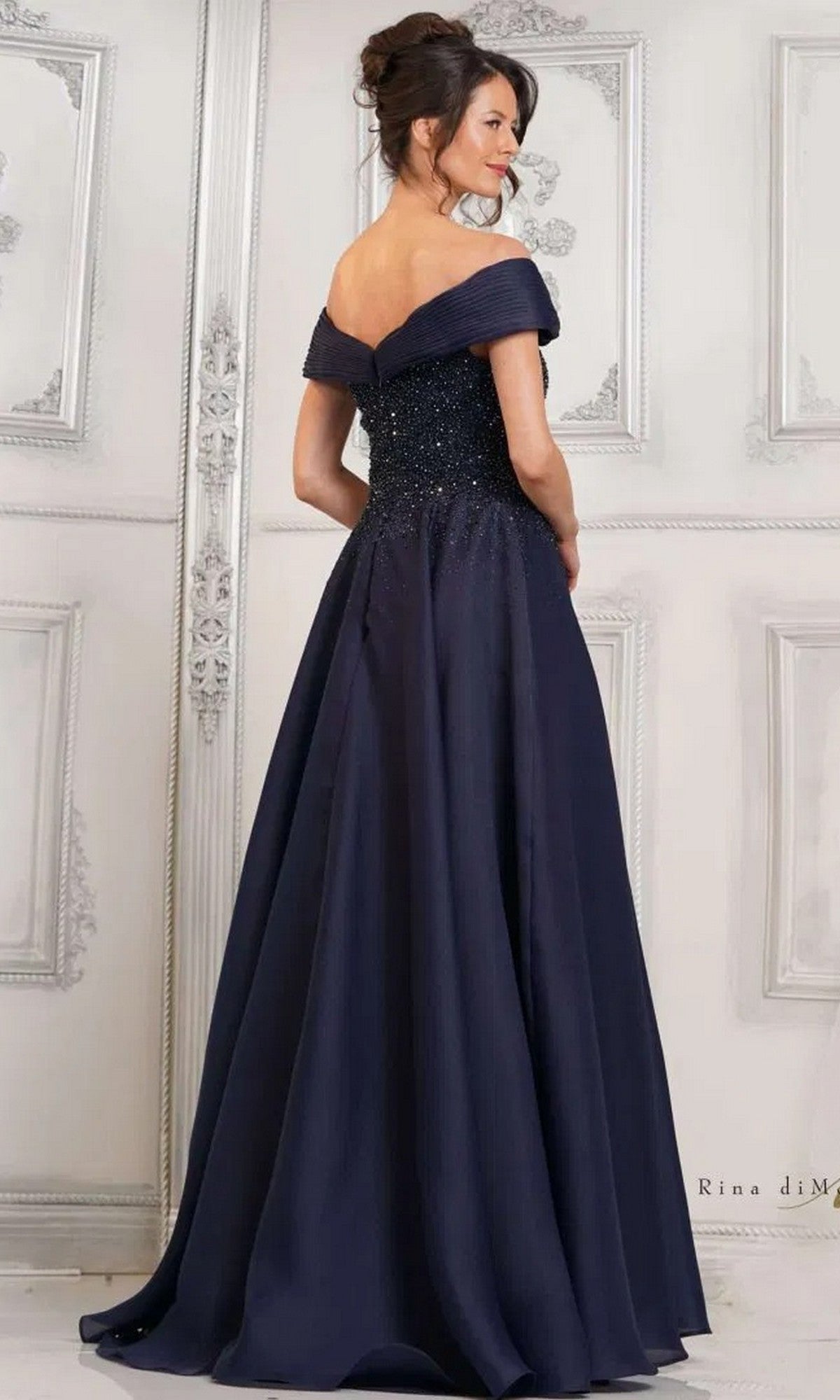 A woman models the Rina Di Montella RD3102 long formal dress, featuring an off-the-shoulder beaded bodice and a flowing, floor-length A-line skirt, posed in an elegant light-colored room.